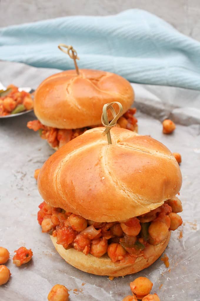 Two vegan sloppy joes with tomato chickpea mixture spilling out of challah buns. Sandwiches are on a countertop with a blue cloth in the back.