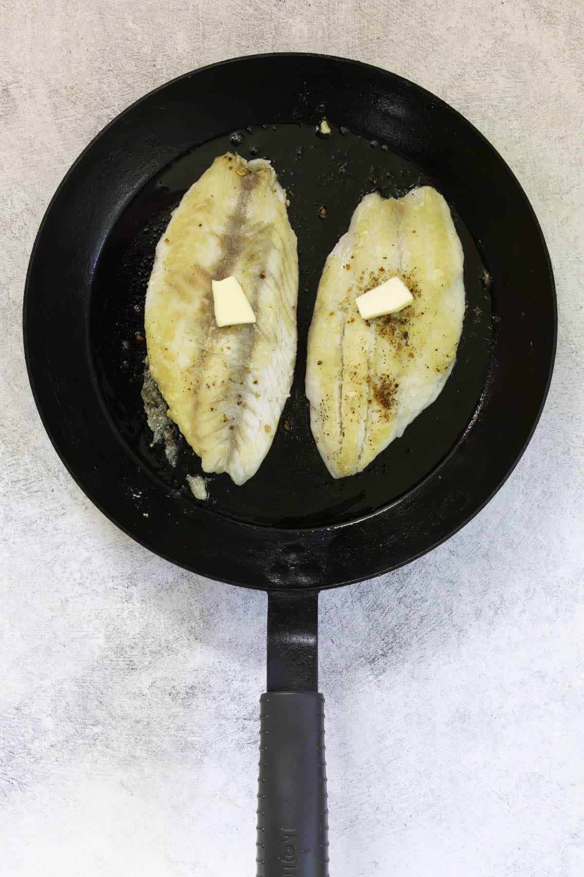 fish fillets topped with butter in hot pan.