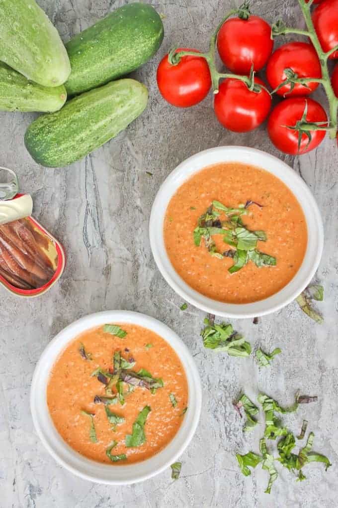Two bowls of summer soup on a granite countertop, surrounded by fresh tomatoes, whole cucumbers, and an open can of anchovies