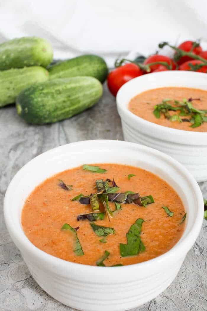 Two bowls of summer soup on a granite countertop, surrounded by fresh tomatoes and whole cucumbers