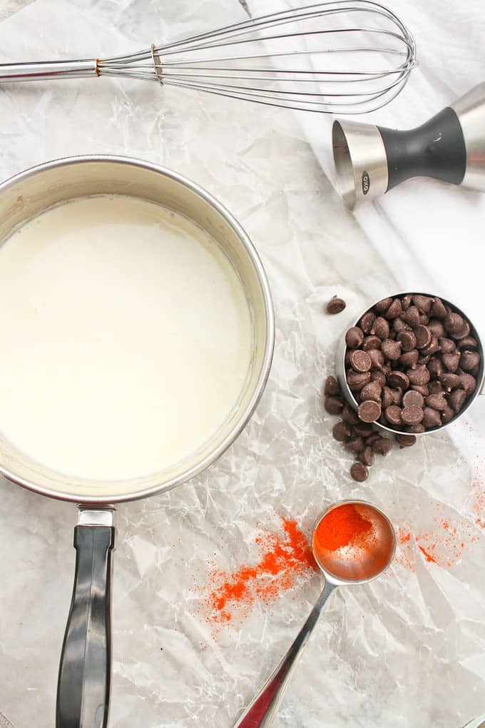 Cream in a saucepan, with chocolate chips, cayenne, a jigger, and a whisk laying on a countertop.