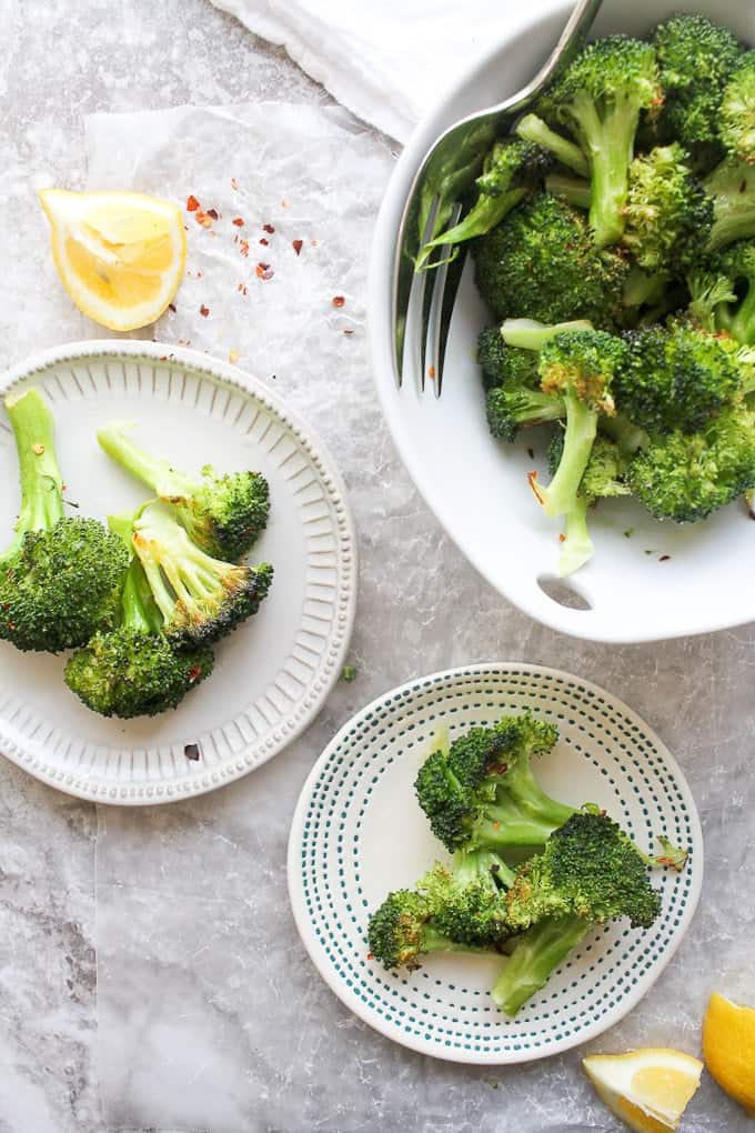 oven-roasted broccoli in a serving bowl and on plates, with lemon and chili flakes on the side