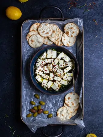 Marinated feta in a bowl on a serving tray with crackers