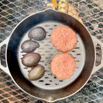 Burger patties and avocado halves in cast-iron pan sitting on campfire grate.