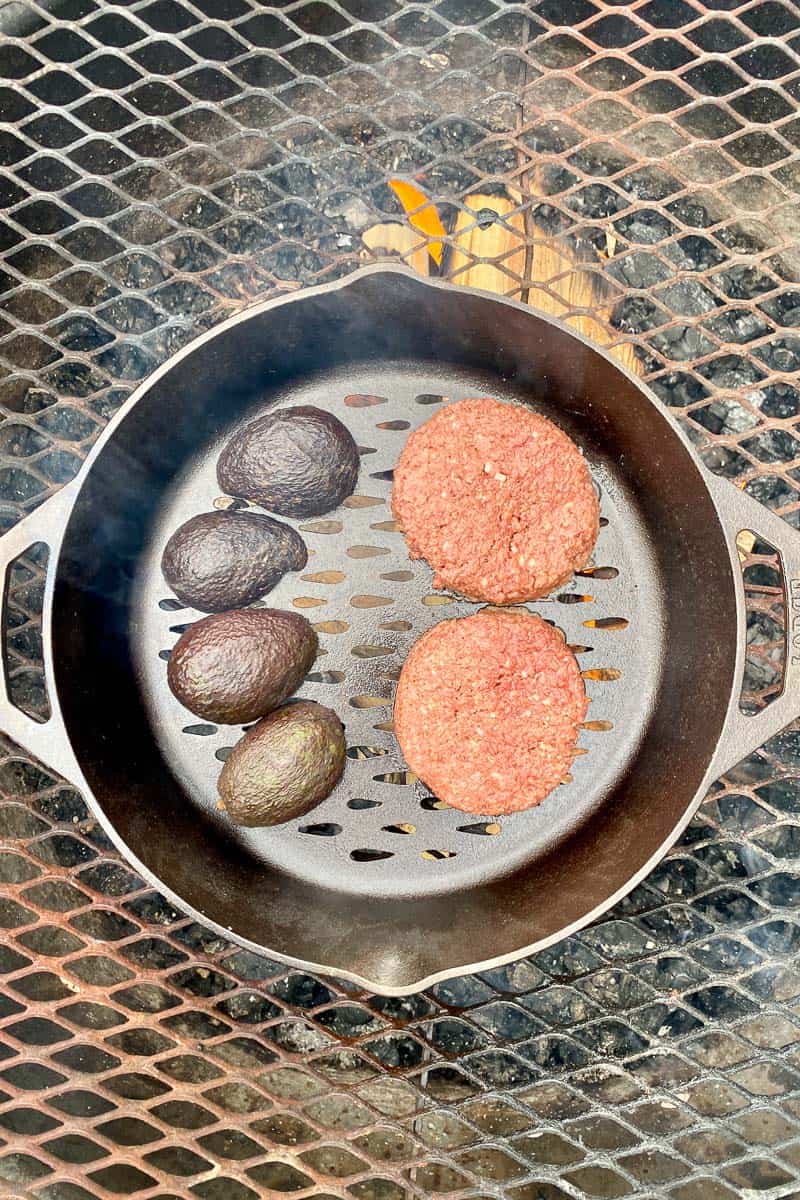 Burger patties and avocado halves in cast-iron pan sitting on campfire grate.