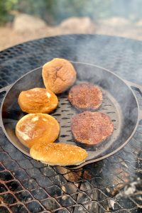 Burger patties and buns sitting in cast-iron pan on campfire grate.