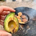 Grilled avocado half with cast-iron pan and campfire in background.