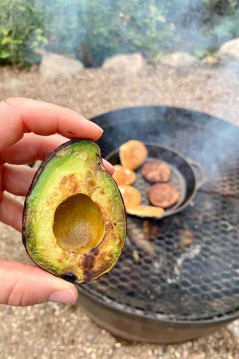 Grilled avocado half with cast-iron pan and campfire in background.