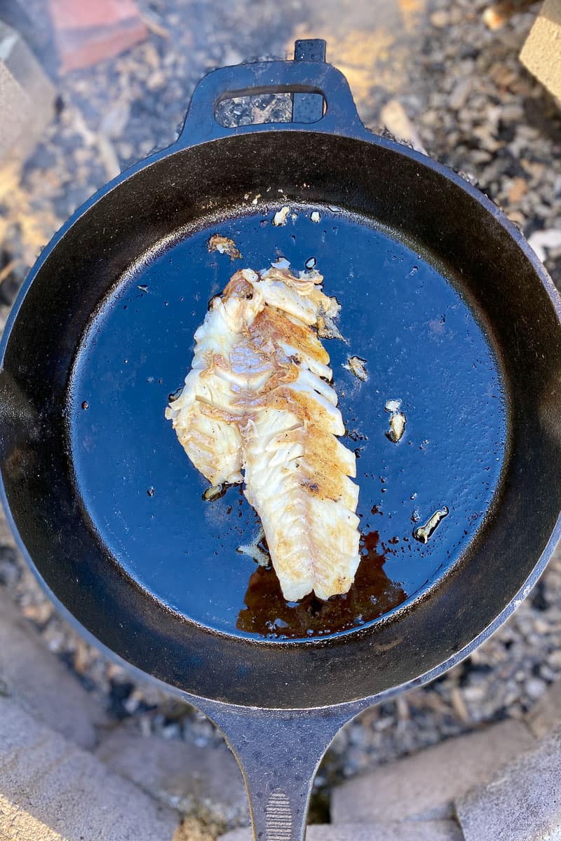 Fish flipped to continue cooking in skillet on campfire grate.