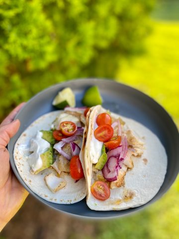 Campfire fish tacos with flour tortillas, tomatoes, avocado, onion and yogurt sauce on plate.