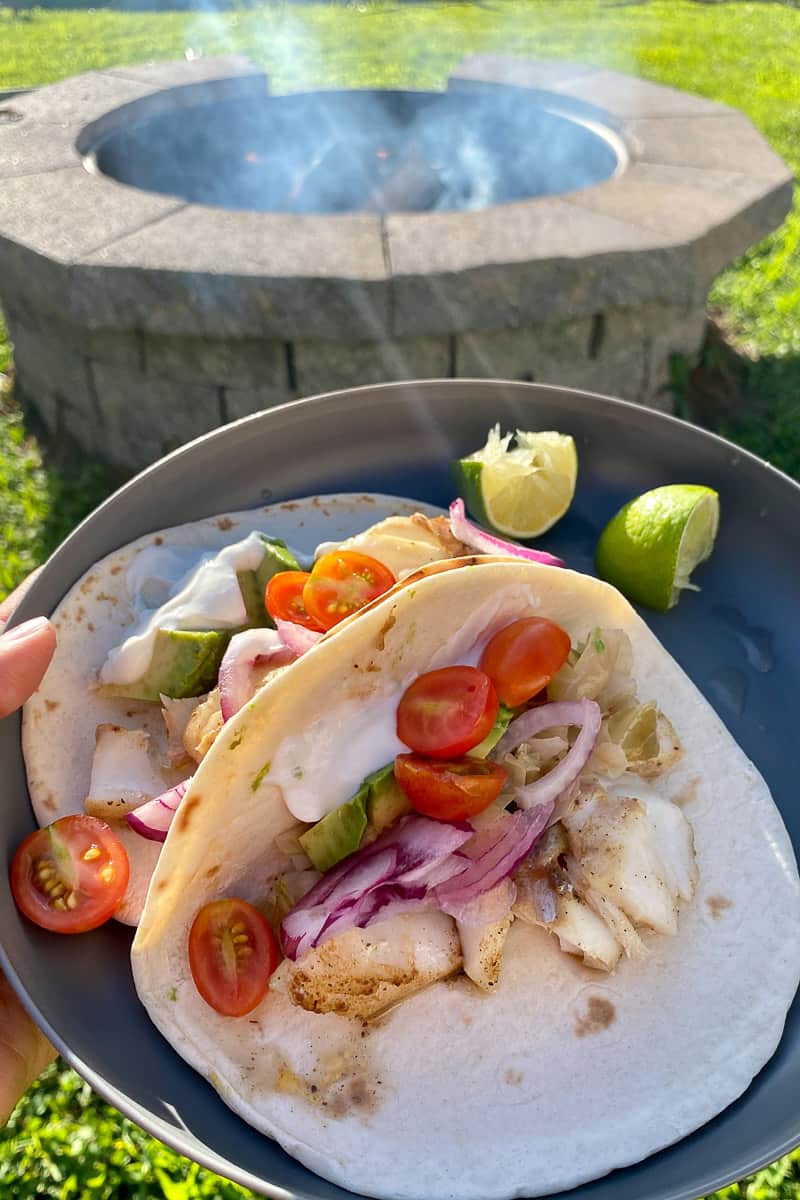 Campfire fish tacos with flour tortillas, tomatoes, avocado, onion and yogurt sauce on plate with fire pit in background.