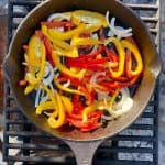 Seasoned peppers and onions in cast-iron pan on campfire grate.