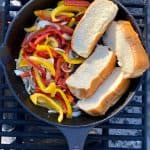 Peppers, onions, and hot dog buns toasting in cast-iron pan on campfire grate.