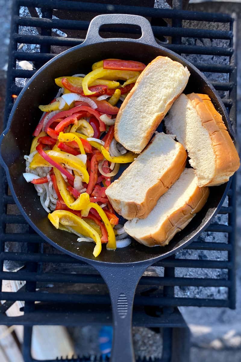 Peppers, onions, and buns toasting in cast-iron pan on campfire grate.