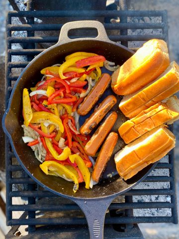 Peppers, onions, hot dogs and buns roasting in cast-iron pan on campfire grate.