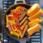 Peppers, onions, hot dogs and buns roasting in cast-iron pan on campfire grate.