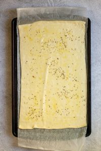 Unrolled pizza dough on baking sheet with oil and herbs spread on top.