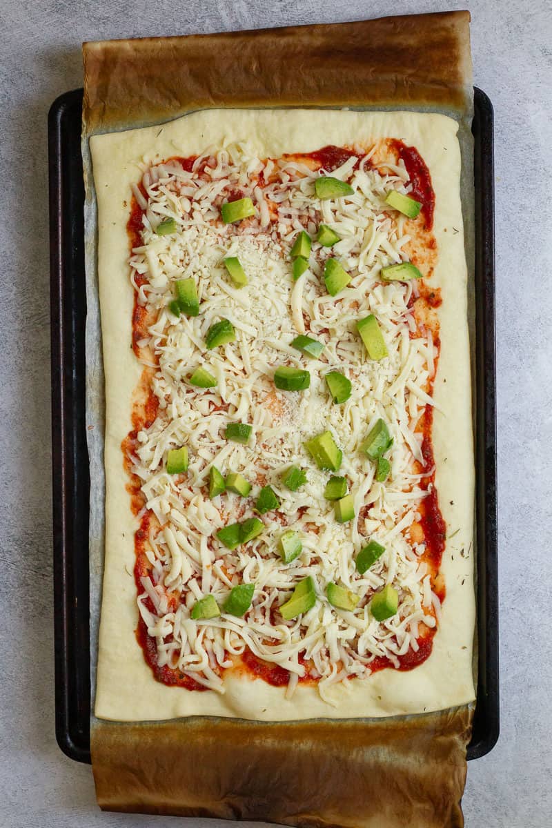 Pizza dough on baking sheet topped with tomato sauce, mozzarella, and avocado.