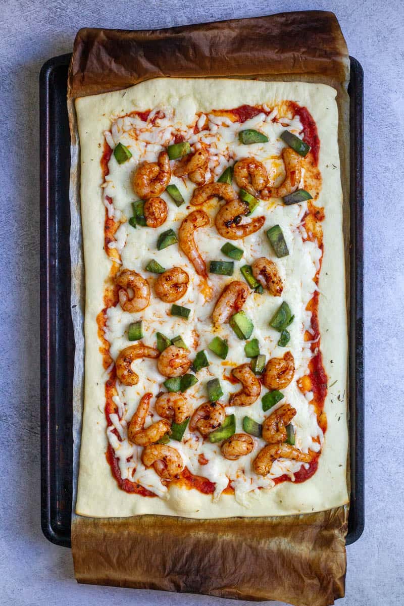 Pizza dough on baking sheet topped with tomato sauce, melted mozzarella, avocado, and spicy shrimp.