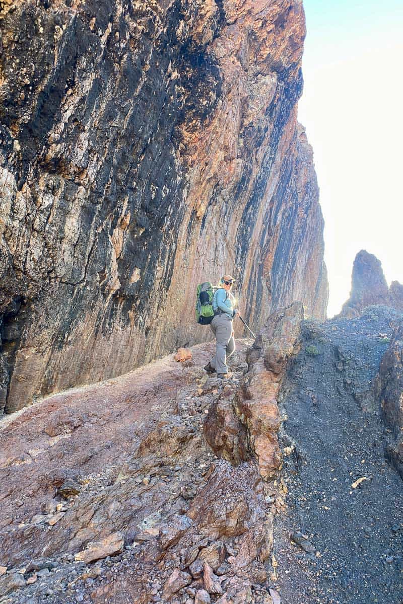 Woman using backpacking pack for petite women as she climbs rocky trail with cliff face rising to the side.