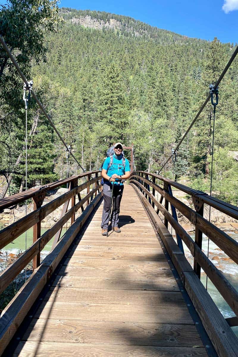 Hiker with trekking pole standing in middle of wooden bridge over creek on Elk Park to Needleton loop.