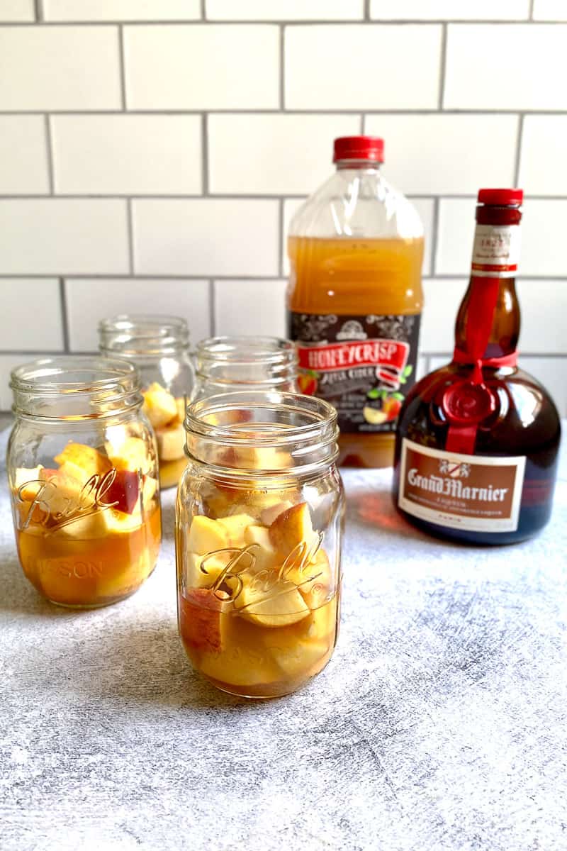 Diced apples in mason jars next to bottles of Grand Marnier and apple cider.
