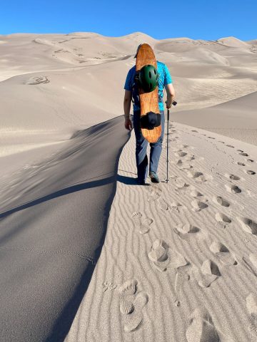 Hiker with sandboard and helmet strapped to his back, leaving footprints as he walks across sand dune.