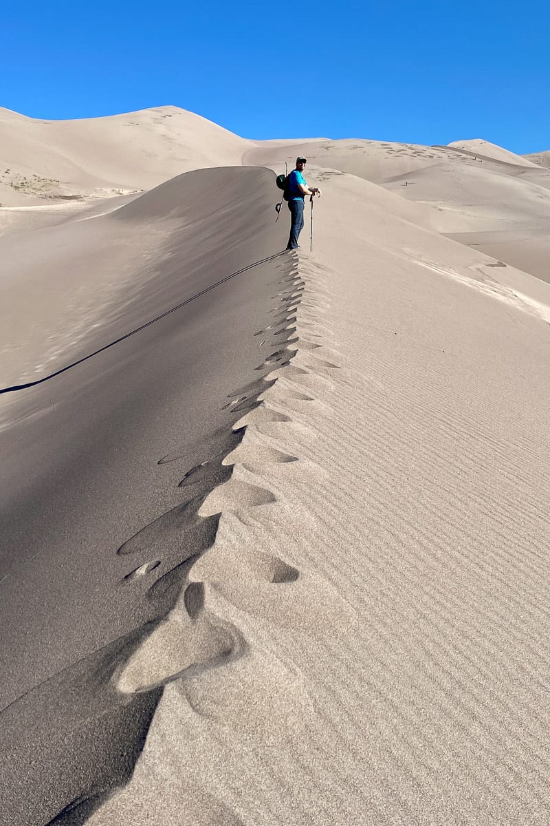 Hiker with sandboard strapped to his back on top of sand dune ridge.