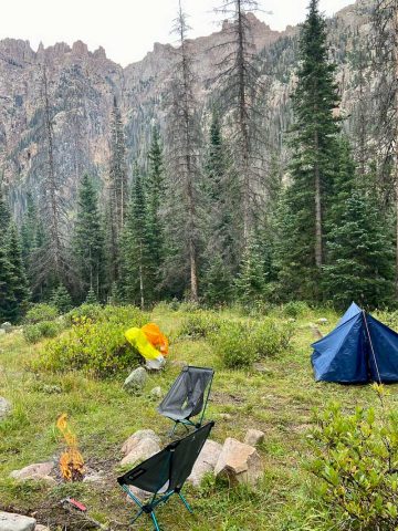 Small campfire, two camp chairs, and small blue tent with tarp over it in field surrounded by trees and mountains.