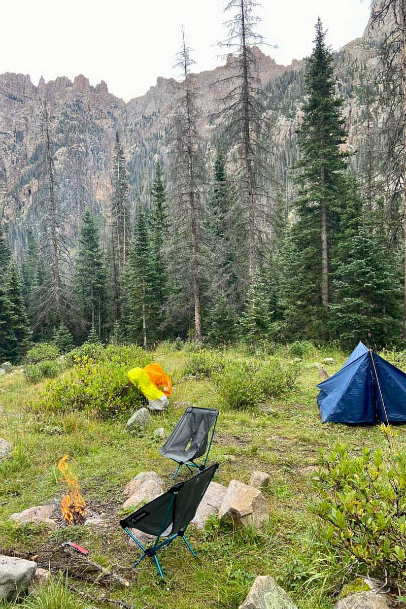 Small campfire surrounded by backpacking gear including camp chairs and small blue tent in field surrounded by trees.