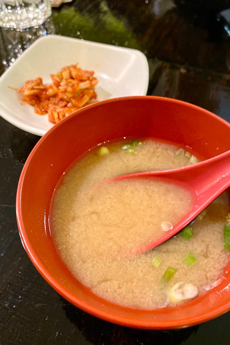 Miso soup in bowl with kimchi on plate in background.