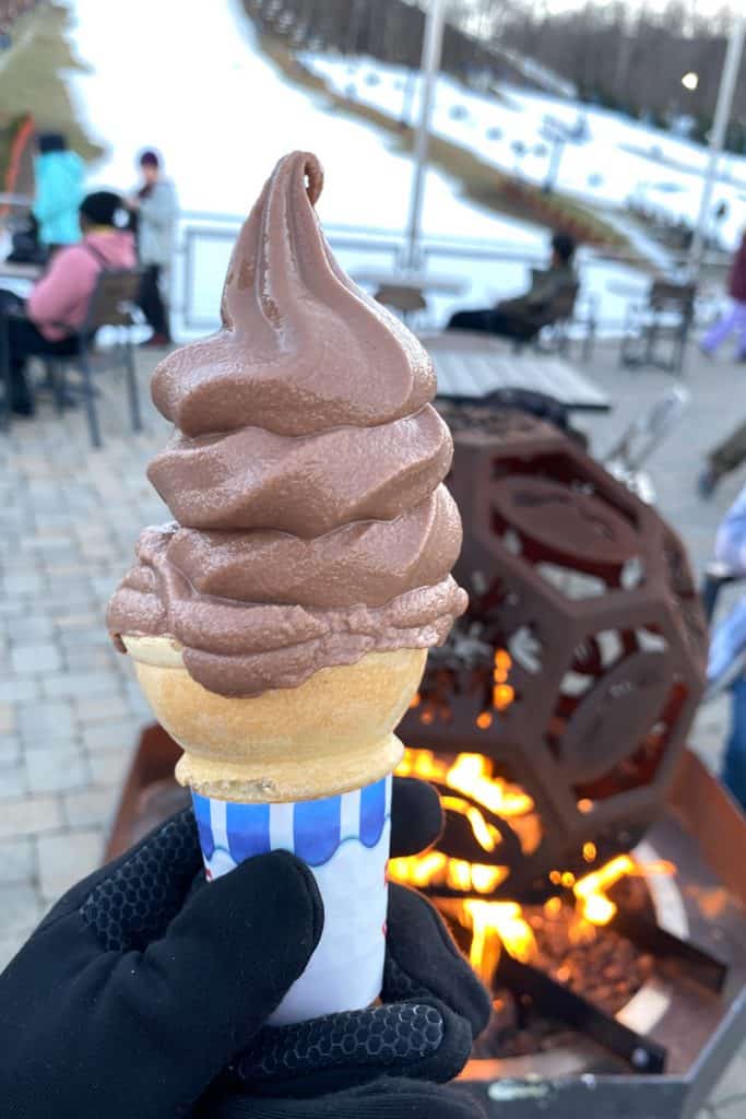 Gloved hand holding chocolate ice cream cone with campfire and ski slopes in background.
