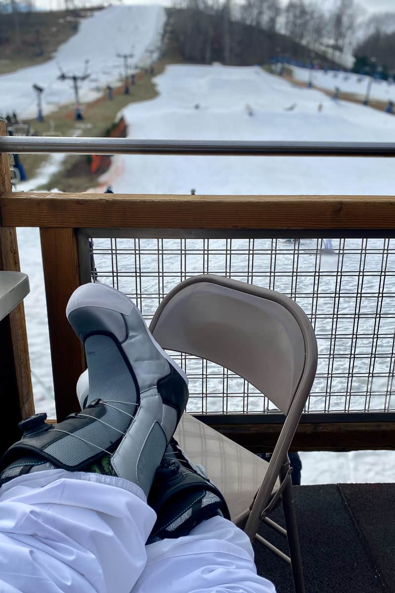 Skier resting booted feet on chair on deck with views of ski run.