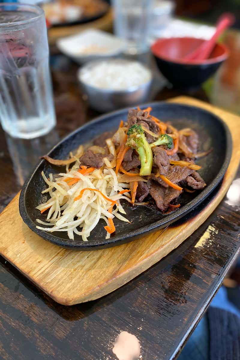 Beef bulgogi with shredded potatoes and cooked veggies served on a cast-iron plate on a wooden board.