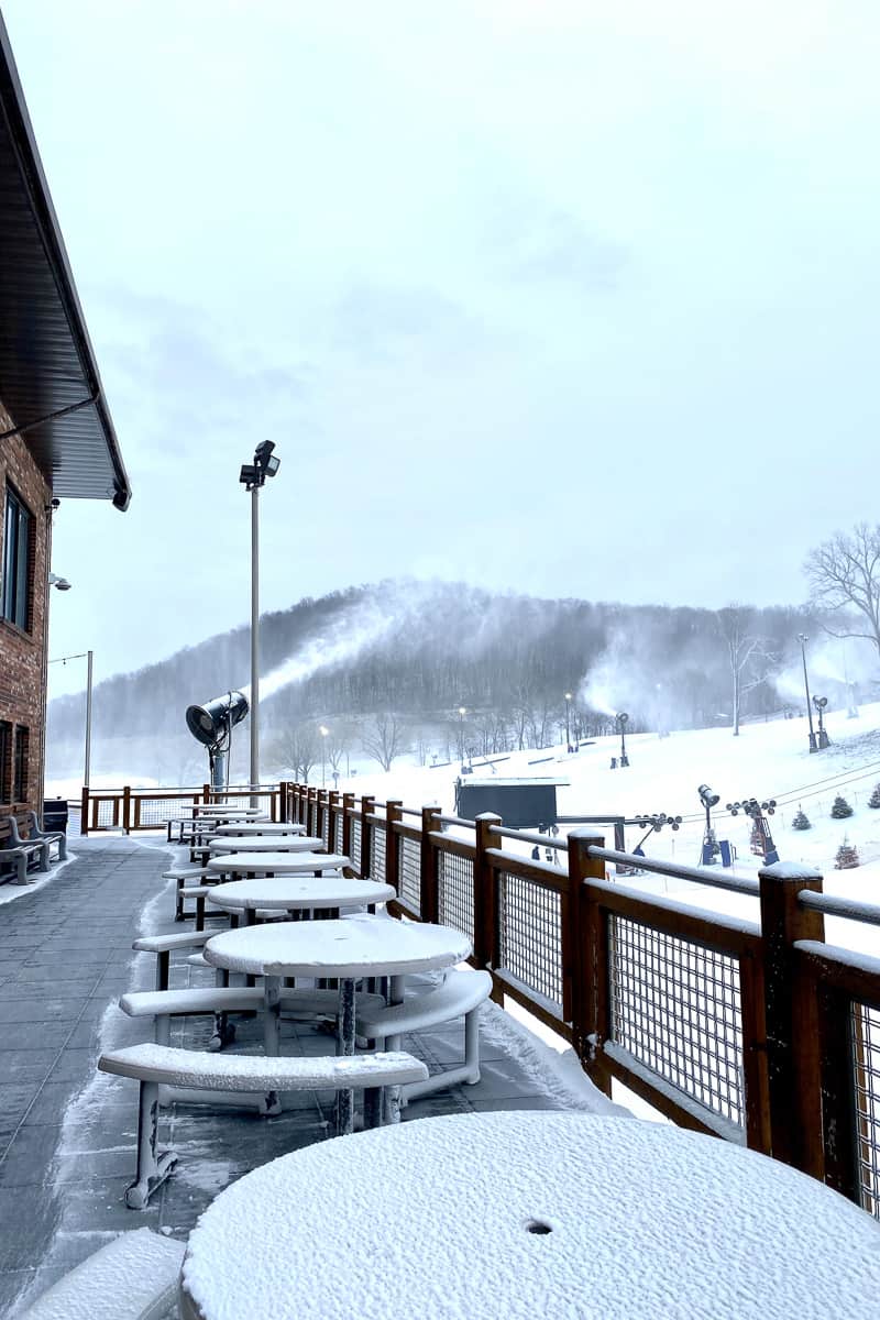Snow covered tables and benches on ski lodge deck.