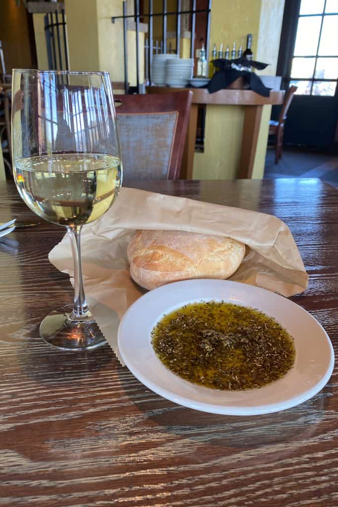 Glass of white wine next to loaf of bread and plate of olive oil and herbs.