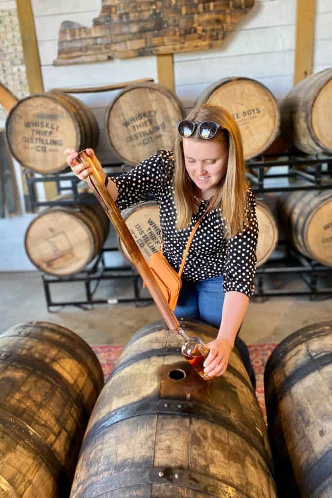 Woman using whiskey thief device to fill glass bottle from barrel.