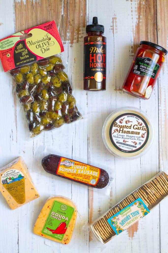 Packages of crackers, cheese, summer sausage, hummus, and olives, and jars of peppers and hot honey.