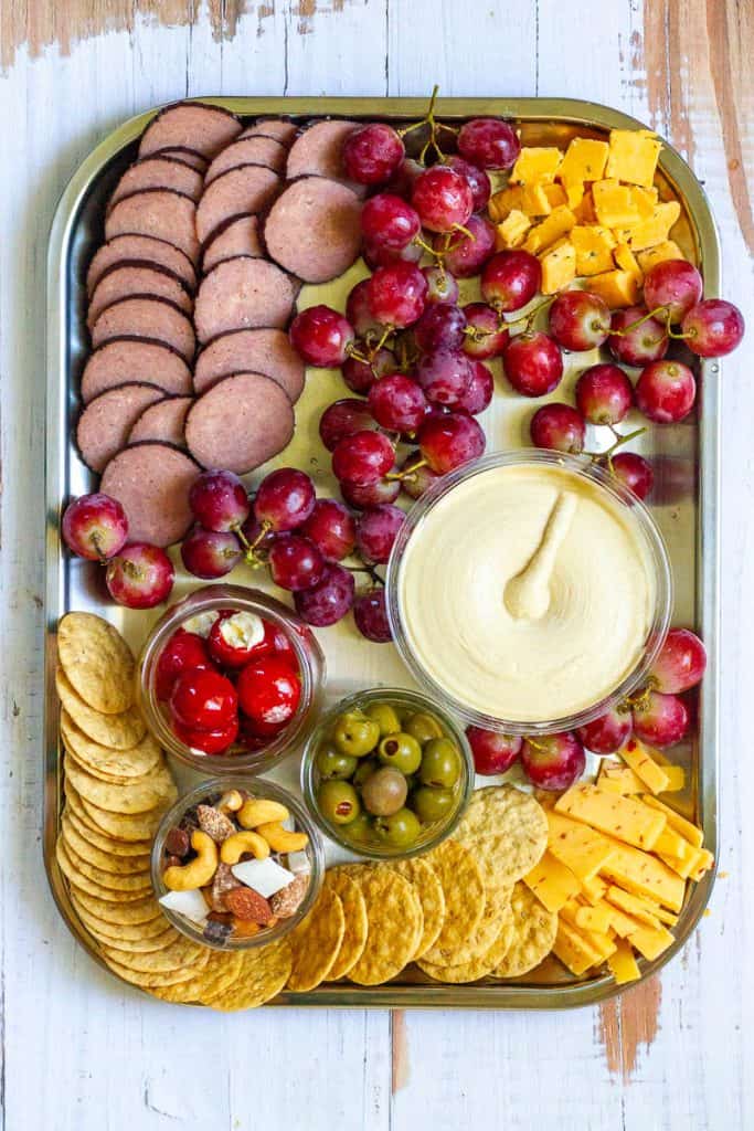 Camping charcuterie board with fruit, crackers, nuts, olives, sausage rounds, cheese and hummus.
