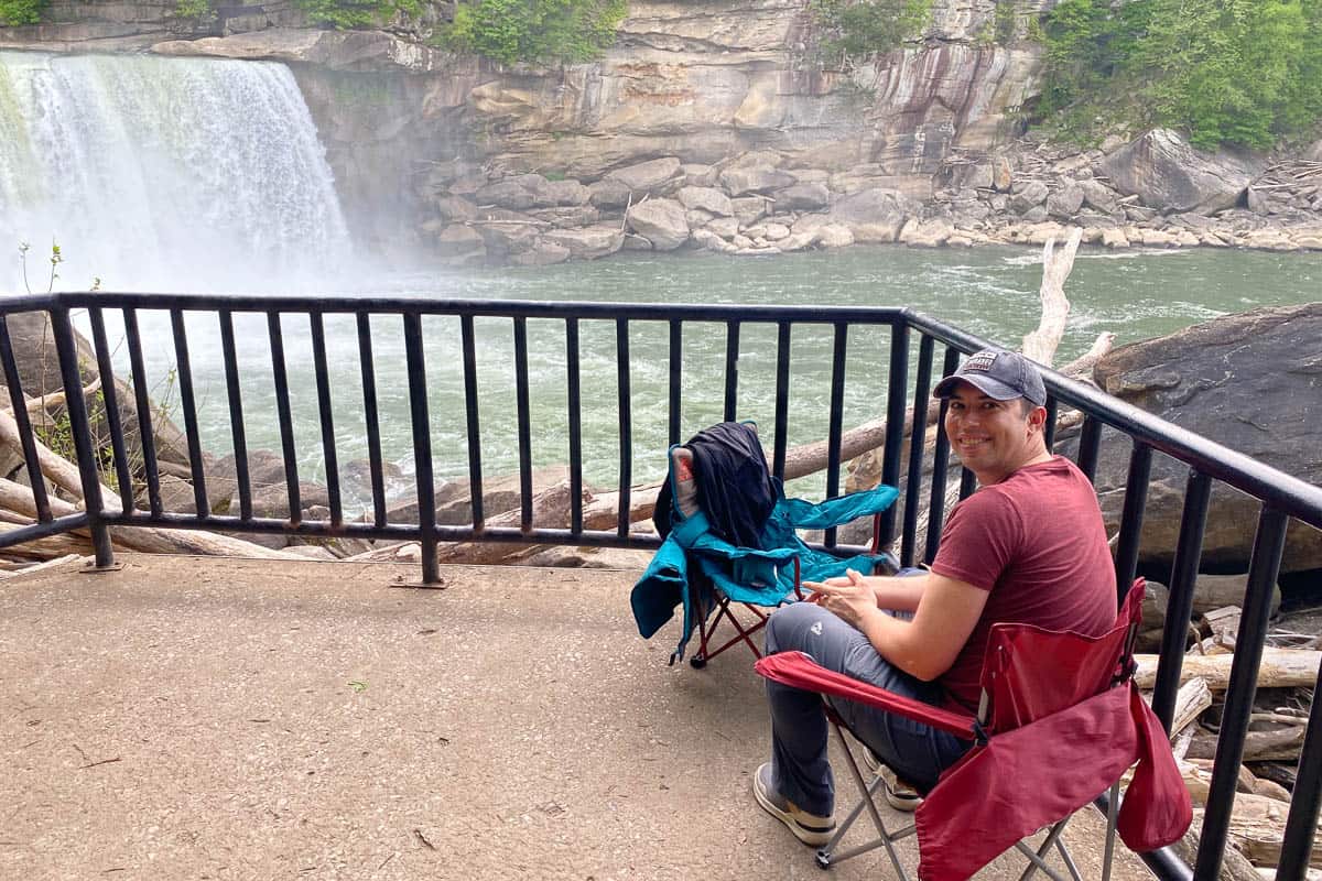 Man seated in camping chair on observation deck near waterfall.