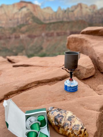 Espresso pods, chocolate croissant, and small gas burner set on red rock.