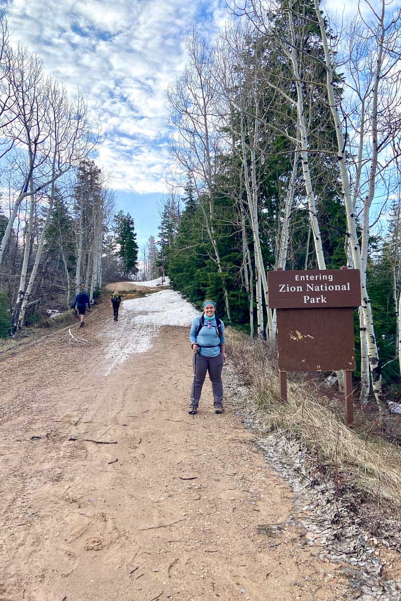 Woman wearing teal sun hoodie for hiking and standing by sign that says "Entering Zion National Park."