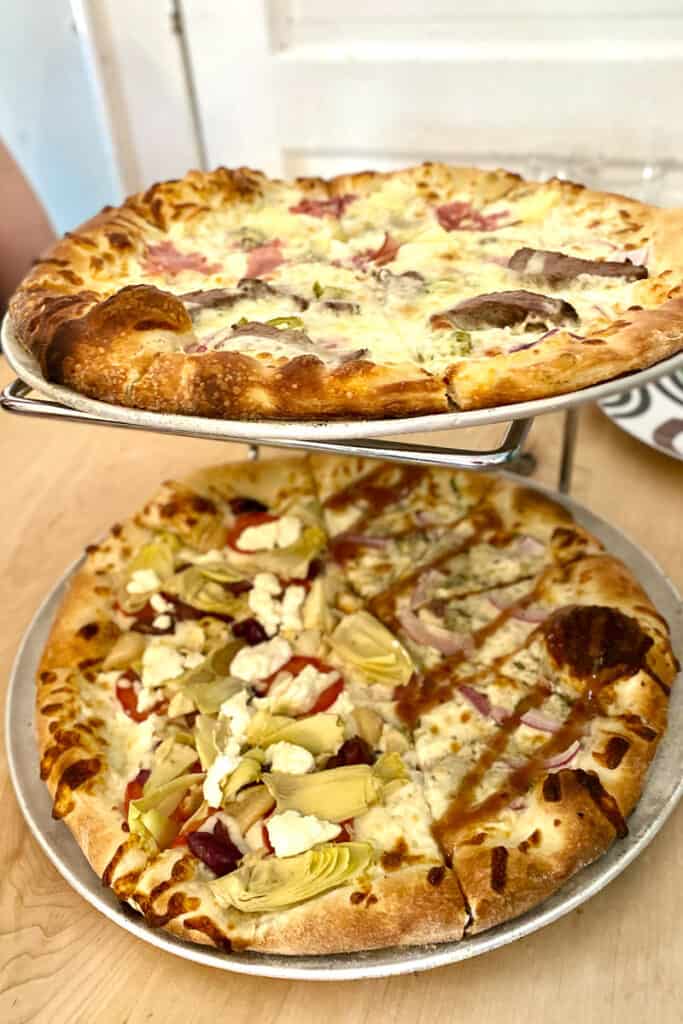 Two pizzas stacked on tiers, one is half topped with fig, jam and gorgonzola and half topped with artichoke and goat cheese; the other is half steak and cheese and half ham and pineapple.