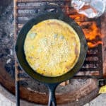 Egg and cheese mixture cooking in cast-iron pan over campfire.