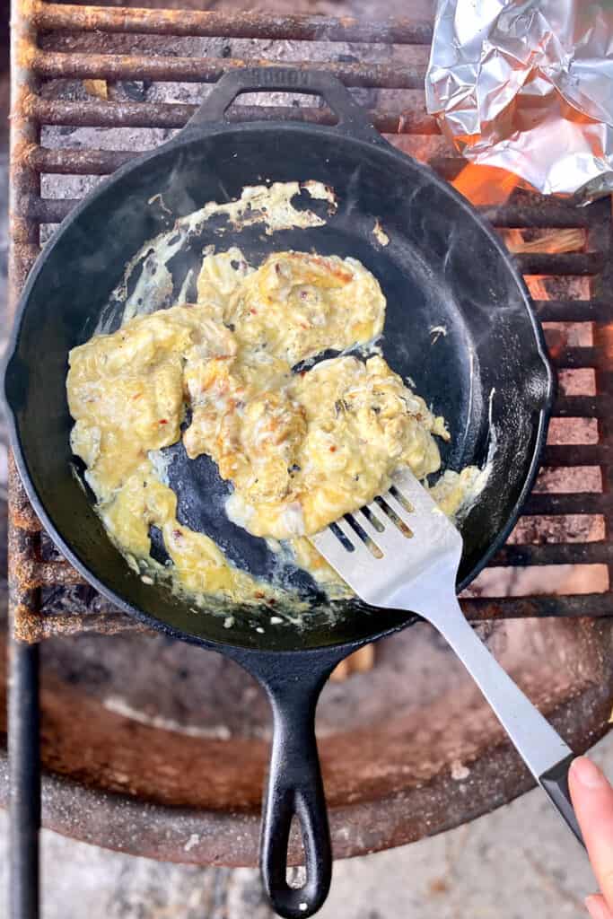 Using spatula to scoop cooked egg and cheese from cast-iron pan for campfire breakfast tacos.