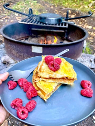 Peanut butter and jelly French toast on plate with raspberries with campfire in background.