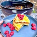 Peanut butter and jelly French toast on plate with raspberries with campfire in background.