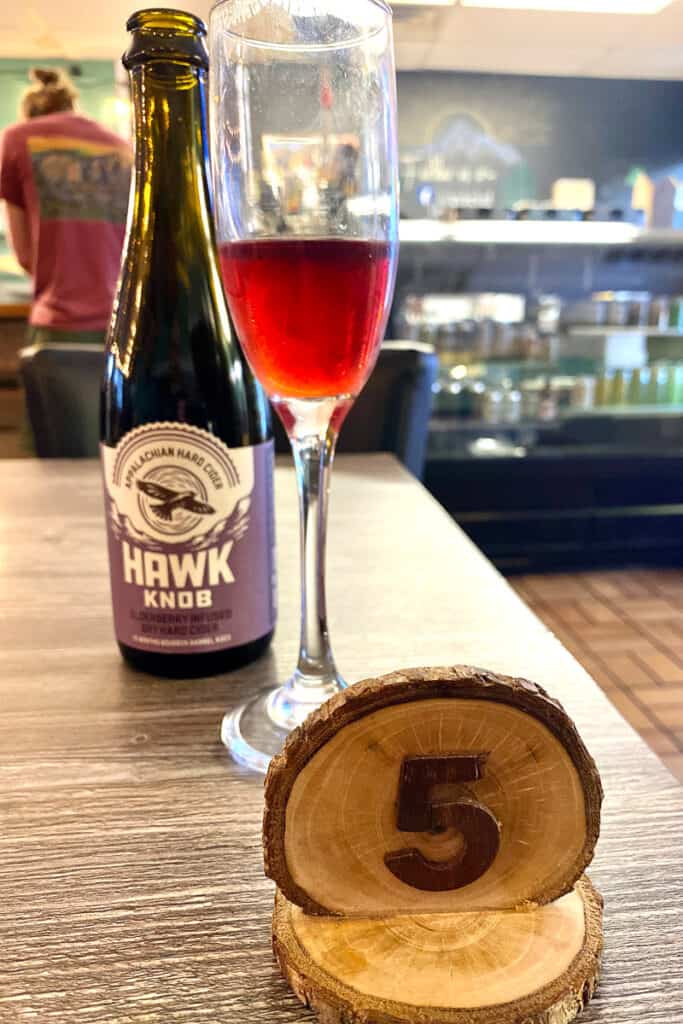 Bottle of Hawk Knob hard cider next to wine glass halfway filled with cider, with wooden medallion number 5 indicating order number.