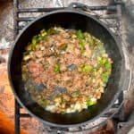 Meat, green peppers and onions in Dutch oven over campfire.