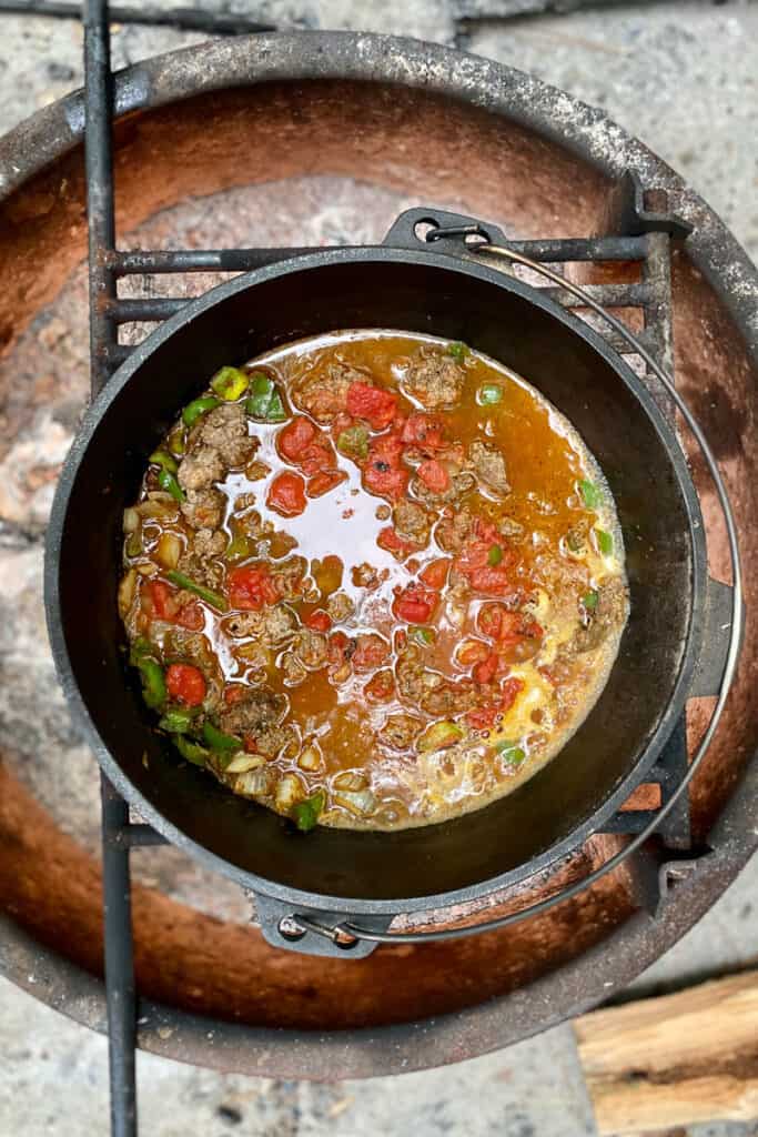 Meat, green peppers tomatoes and onions in broth in Dutch oven over campfire.